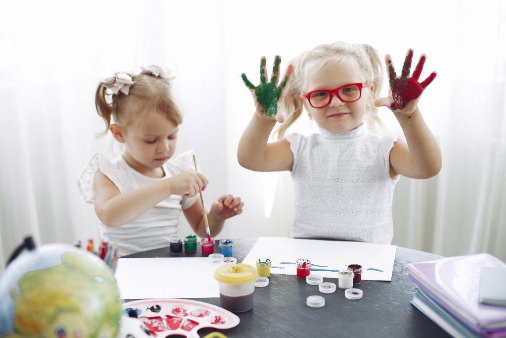 two little girls play with paint at a table with their fingers and brushes.