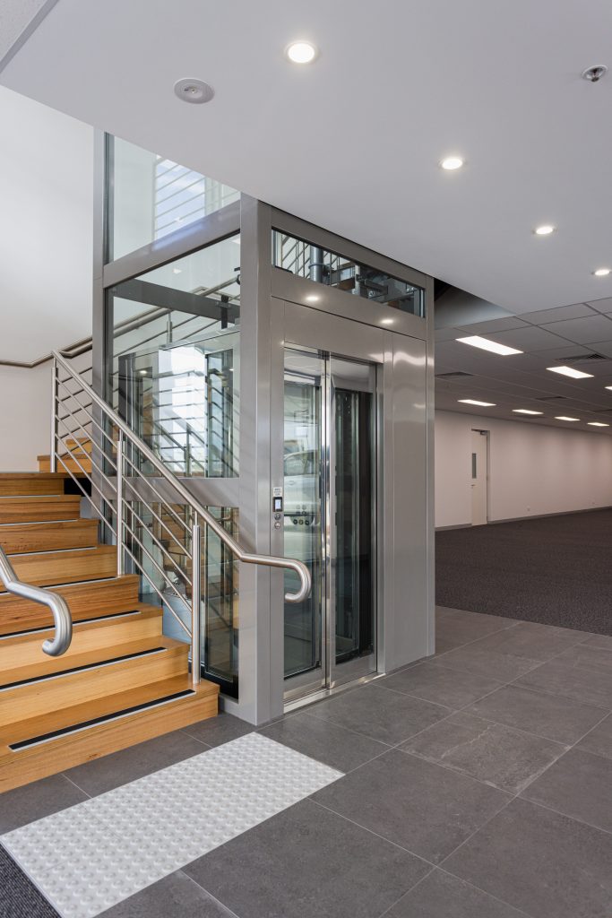 grey lift with clear doors is next to a wooden staircase inside a commercial building.