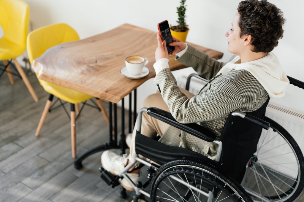 a person who lives with a disability is sitting in a wheelchair at a table drinking coffee while on her phone.