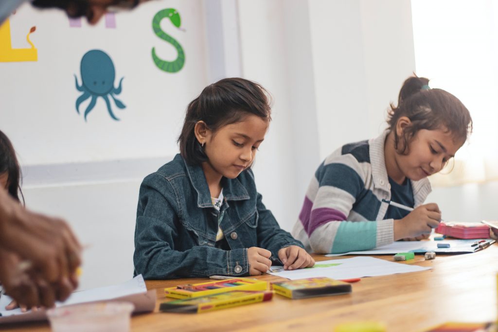 children sit at a school desk while doing arts and crafts.