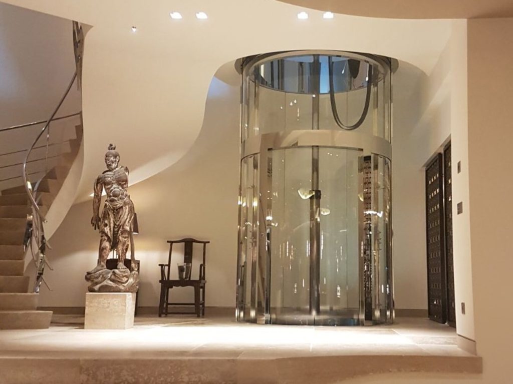 Modern glass lift beside ancient statue and stairs.