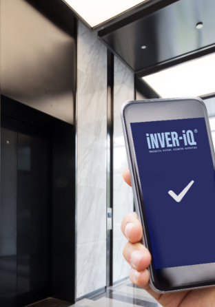 A hand holding a smartphone displaying a screen with the text "INVER-iQ" and a checkmark. The background shows a modern lobby area with a closed elevator door and marble walls, as well as large windows allowing natural light into the space.