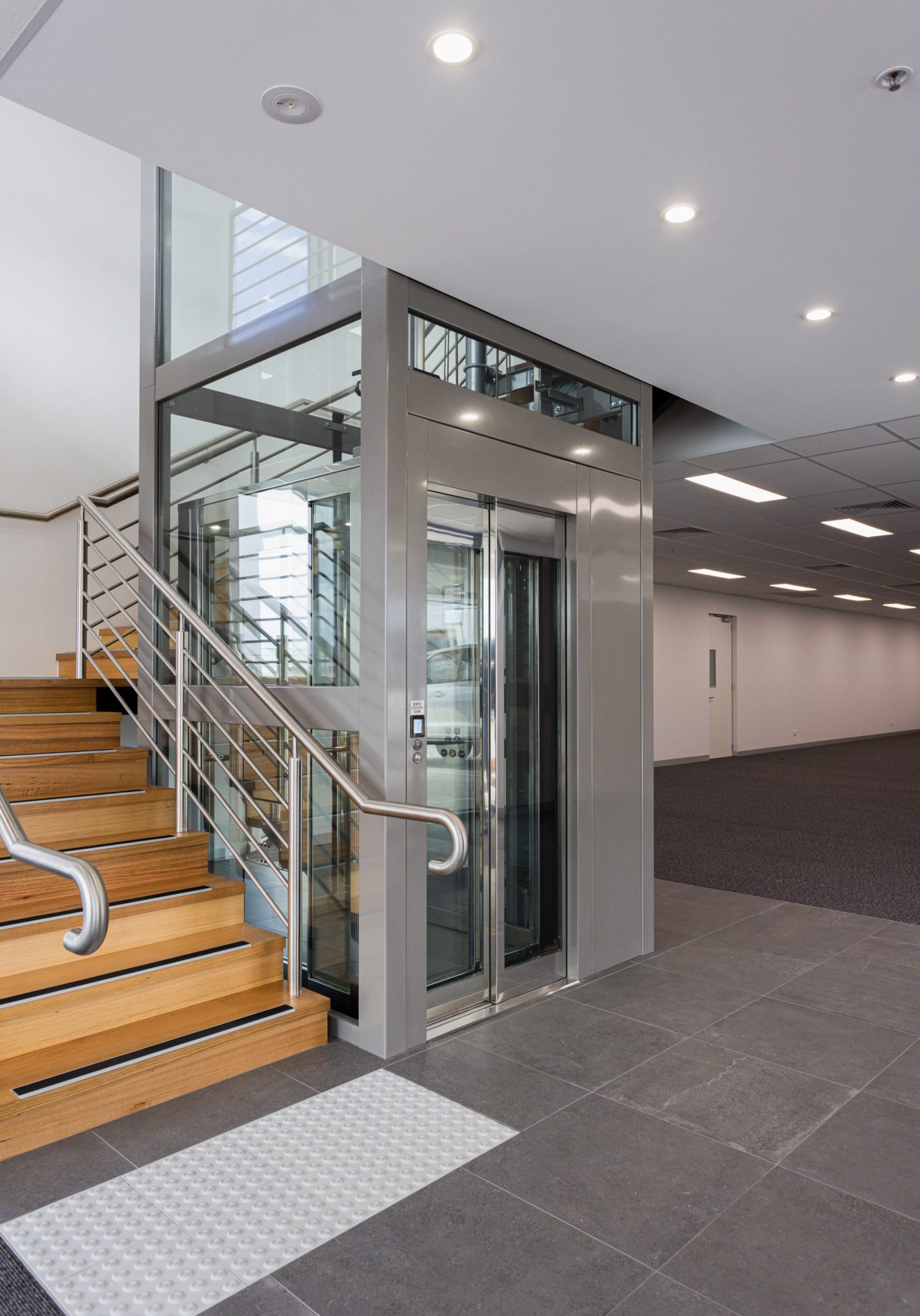 grey lift with clear doors is next to a wooden staircase inside a commercial building.